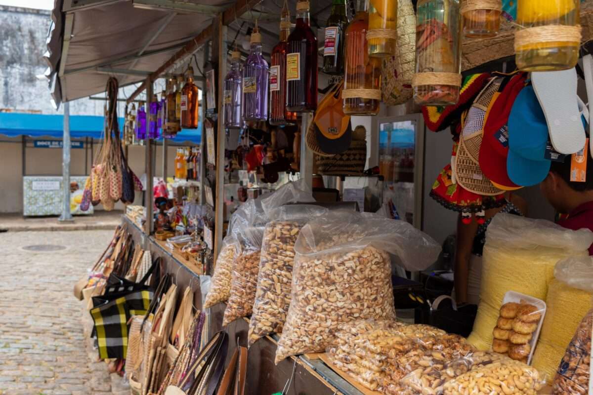 Especiarias da culinária nordestina. Na imagem: barracas com castanhas do Pará na bancada, bolsas de palha e plástico no tampo externo; cachaças e garrafas com conservas estão penduradas em cima e do lado de dentro, diferentes acessórios de vestuário e grãos.